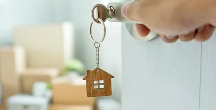 A hand holds a key with a house-shaped keychain inserted into a door, with moving boxes blurred in the background.