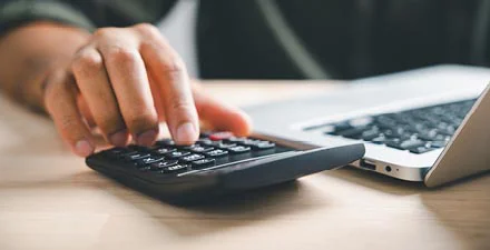 Person using a calculator next to a laptop on a light wooden desk.
