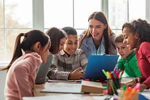 Teacher and diverse group of students smiling and looking at a laptop together in a bright classroom.
