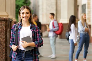 Smiling female student holding a tablet and backpack, standing outside a university building with other students in the background.