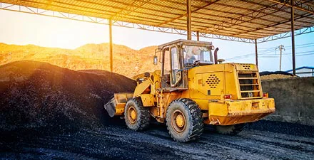 Yellow front loader moving piles of black gravel under a large metal canopy at sunset.