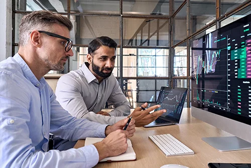 Two men analyzing financial charts on computer screens in an office with many windows..