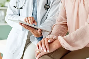 Doctor reviewing medical information on a tablet while a patient sits nearby with hands folded.