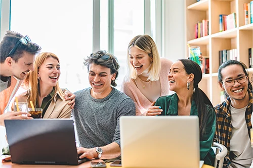 Group of college students on two laptops smiling in library together.