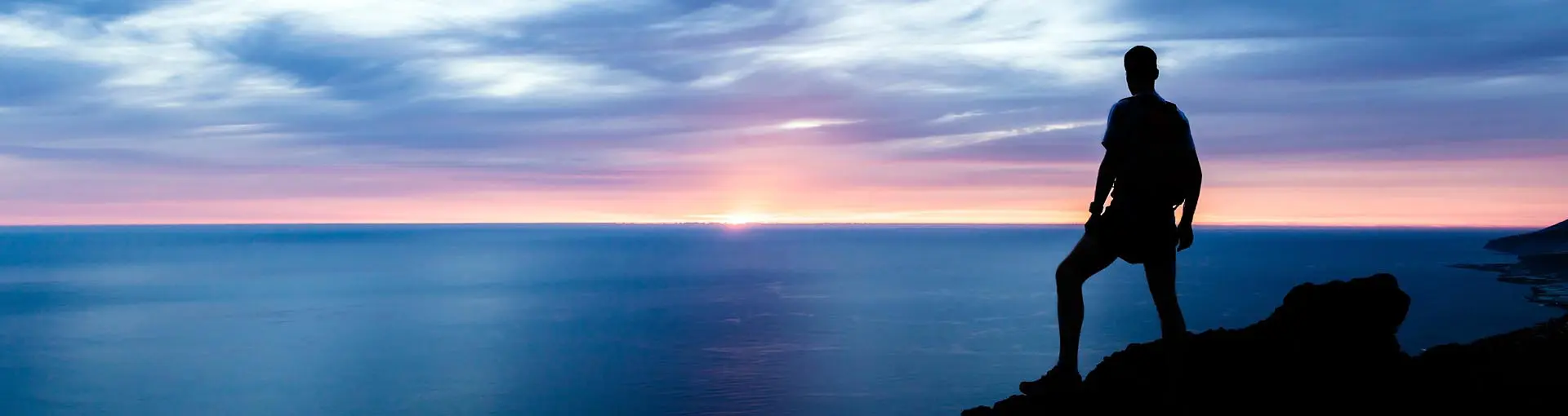 Silhouette of a person standing on a cliff overlooking ocean at sunset.