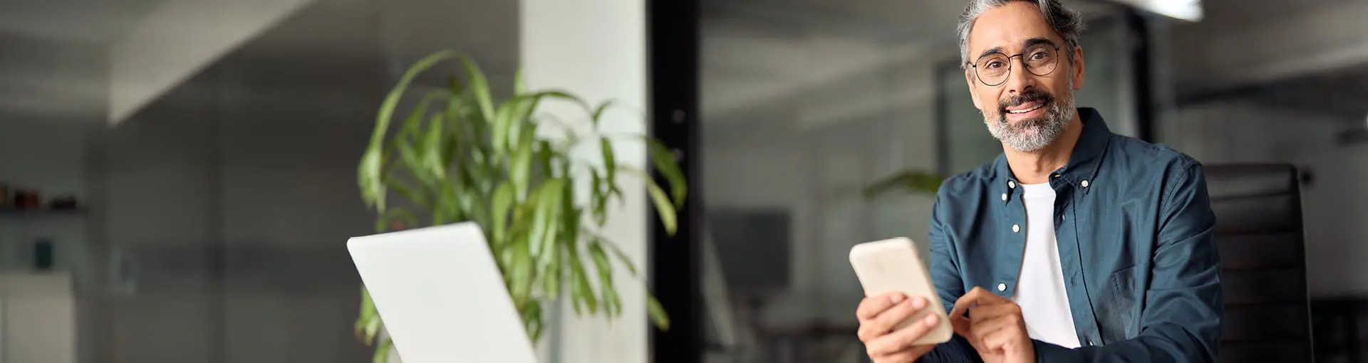 Smiling man with glasses using a smart phone at a desk with a laptop and plant in the background.