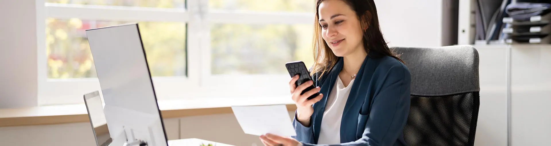 Woman sitting at a desk holding a phone and looking at a document with a computer in front of her.