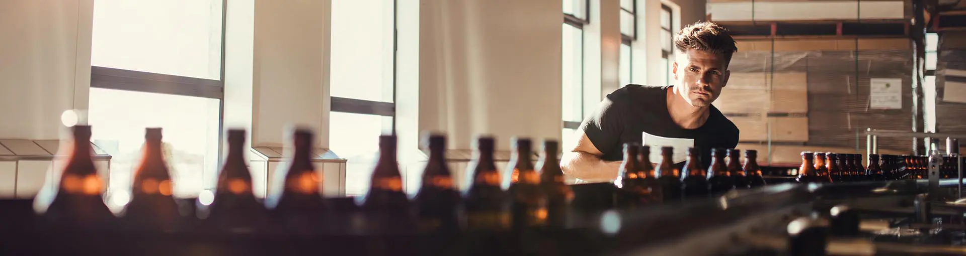 Man inspecting brown glass bottles on a conveyor belt in a sunlit beverage manufacturing facility.