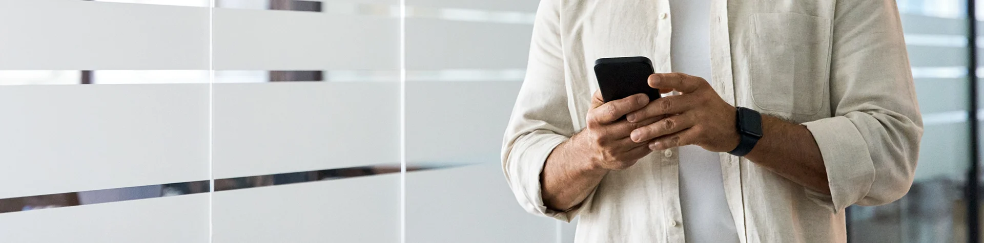 Man standing by window holding his mobile phone in tan dress shirt.