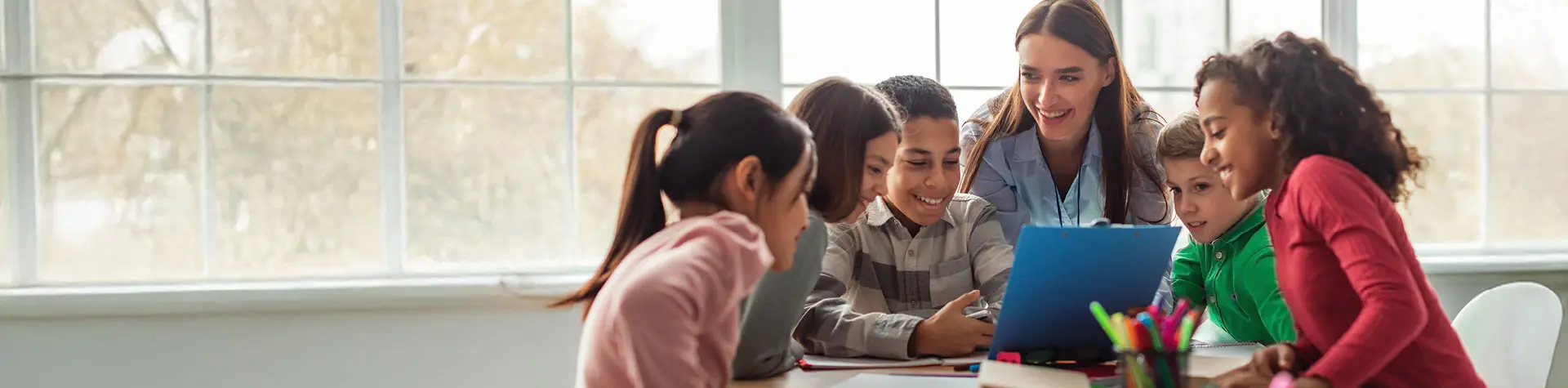 Smiling teacher and diverse group of students gathered around a laptop in a bright classroom.