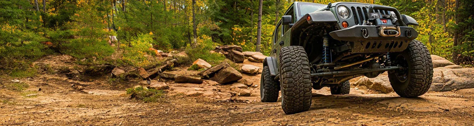 Jeep Wrangler driving off-road through a rocky forest trail surrounded by trees and bushes.