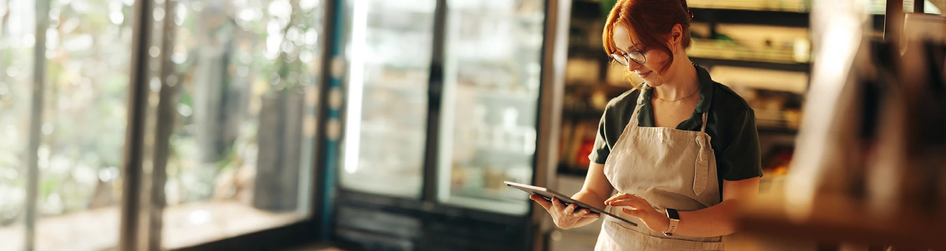 Female owner with apron on inside of open concept shop with windows working on a tablet.
