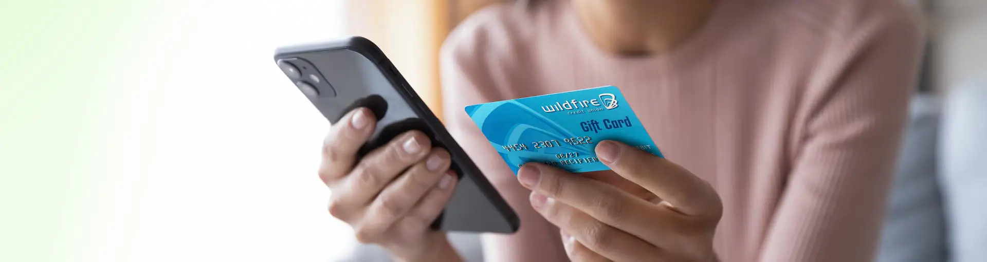 Woman in salmon shirt holding a smartphone and a blue Wildfire Credit Union gift card.