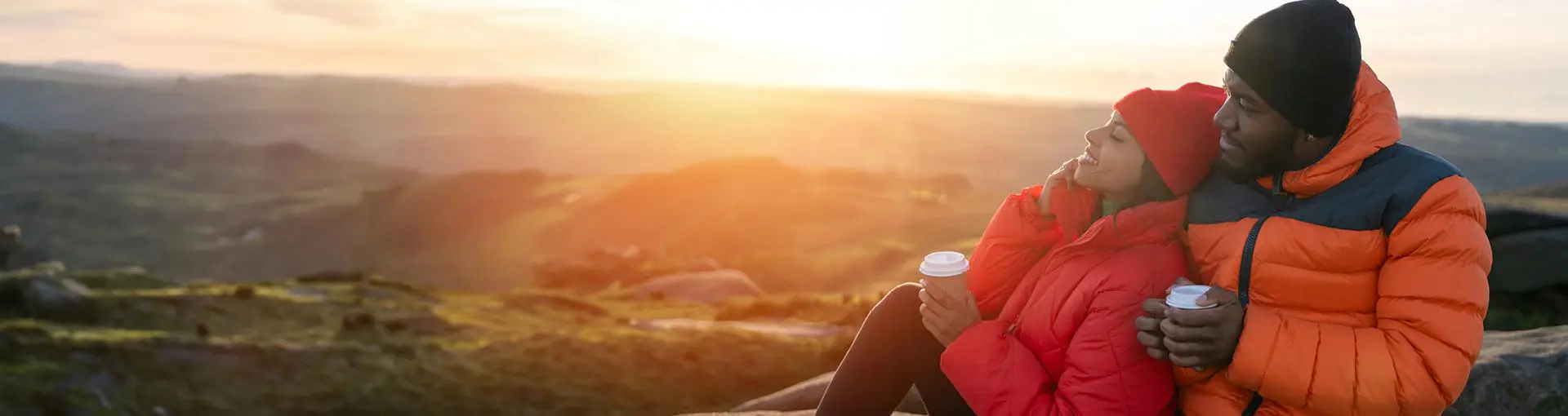 A couple sitting on top of a mountain drinking coffee looking over the scenery in winter coats.