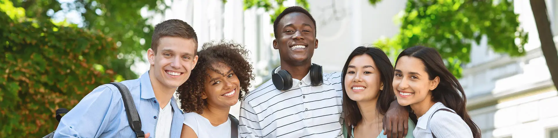 A group of young college students gathered outside university on sunny day with trees in background. .