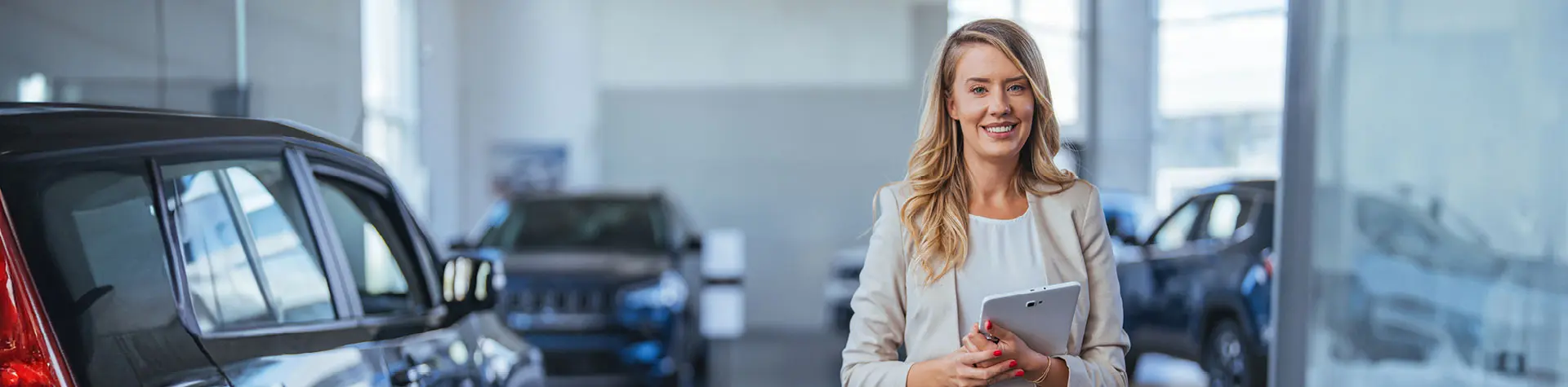 Smiling woman holding a tablet in a car showroom, standing next to a black SUV with vehicles in the background.