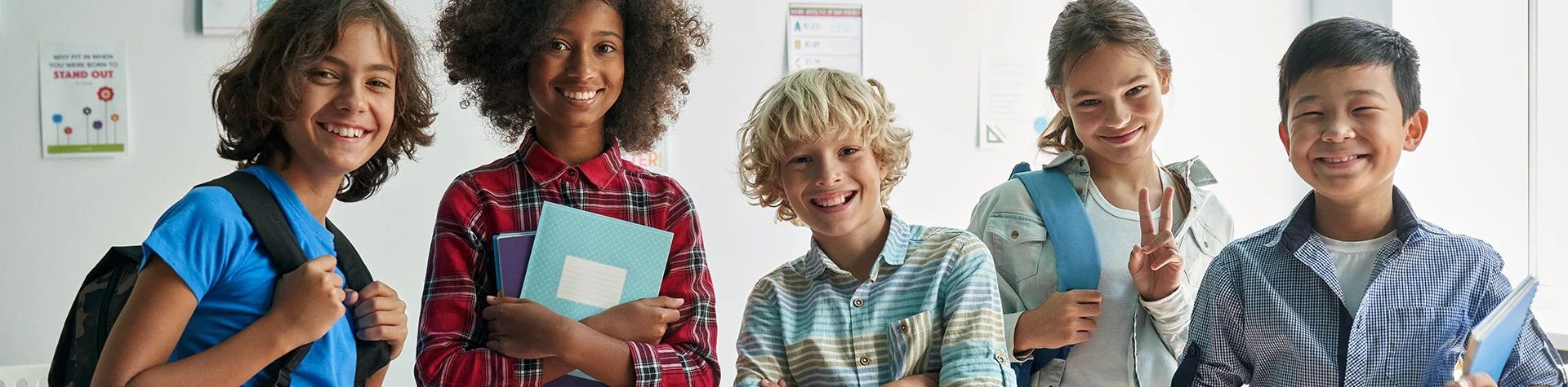 Five smiling school children standing together indoors with backpacks and notebooks, ready for school.