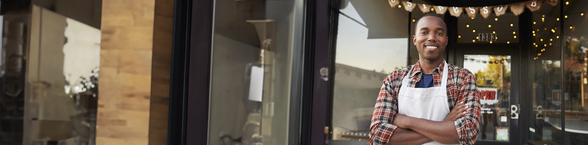 Man in flannel shirt and apron standing outside of downtown business window smiling.