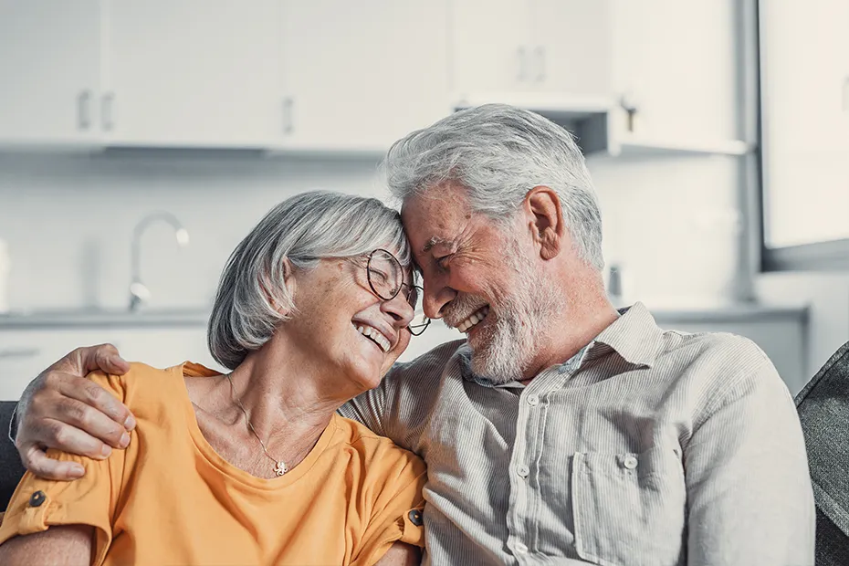 Elder couple in white house laughing and hugging with foreheads on each others.