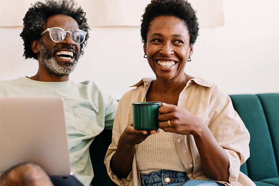 Smiling couple sitting on green couch with laptop and coffee in hand.