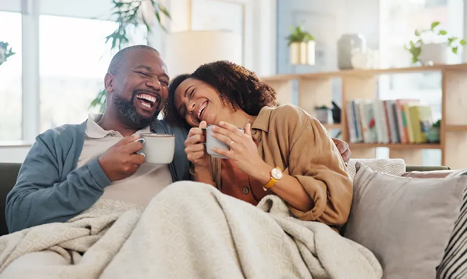 Couple laughing in house while drinking coffee on a couch with a cozy blanket.